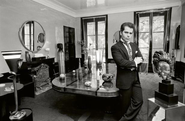 Karl Lagerfeld posiert in einem dunklen Anzug in einem eleganten Raum mit Kunstobjekten.
