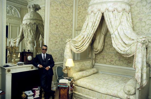 Karl Lagerfeld posiert in einem eleganten Raum mit einem Himmelbett und floralen Tapeten.