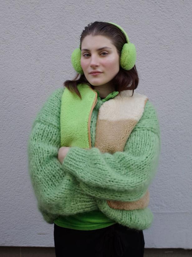 Eine Frau mit grünen Ohrenschützern und einem grünen Strickpullover vor einer grauen Wand.