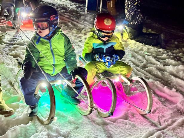 Zwei Kinder beim Nachtrodeln auf Schlitten mit grüner und pinker Beleuchtung auf einer beschneiten Rodelstrecke 