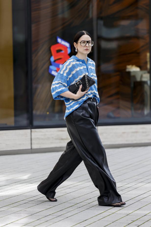 Eine Frau mit Brille, blau-weiß gestreiftem Oberteil und schwarzer Hose geht auf dem Bürgersteig.