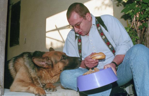 Ein Mann mit Hosenträgern füttert einen Schäferhund mit einem Brötchen.