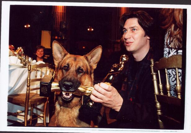 Ein Mann sitzt an einem Tisch mit seinem Schäferhund, der eine goldene Statue im Maul hält.