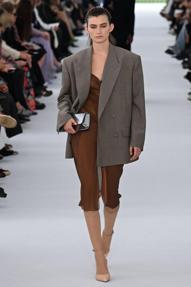 Eine Frau in einem braunen Kleid und übergroßem Blazer auf dem Laufsteg.