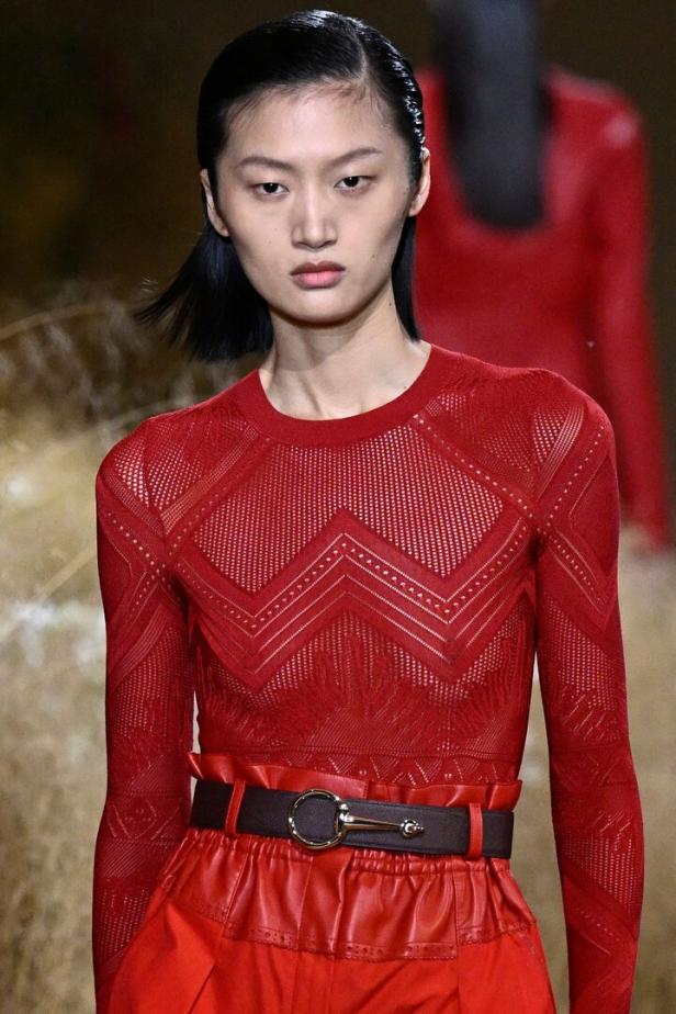 Eine Frau mit dunklen Haaren trägt ein rotes Outfit mit einem gemusterten Oberteil und einem Gürtel.