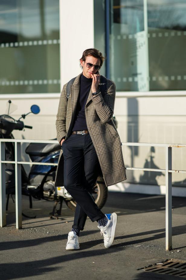 Ein Mann mit Sonnenbrille, Mantel und Turnschuhen telefoniert auf der Straße.