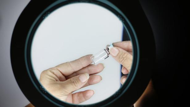 Eine Person hält eine kleine Phiole mit einer klaren Flüssigkeit unter einer Lupe.
