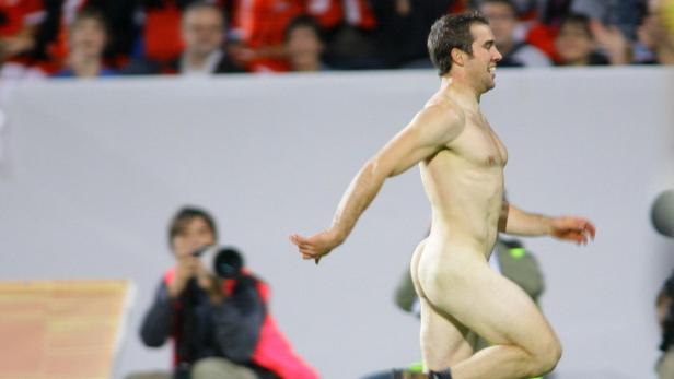 Ein nackter Mann rennt über ein grünes Feld vor einer Zuschauertribüne.