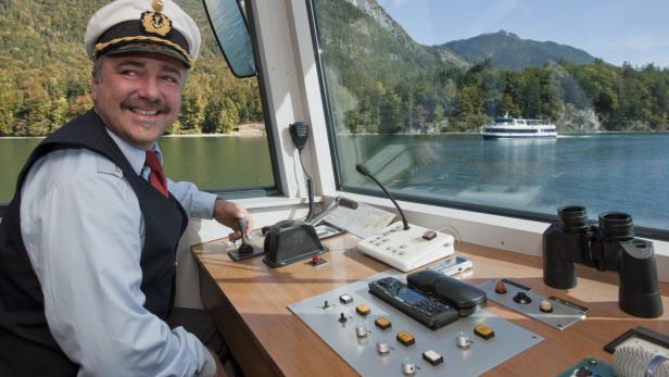 Ein Kapitän steuert ein Schiff auf einem See mit Bergen im Hintergrund.