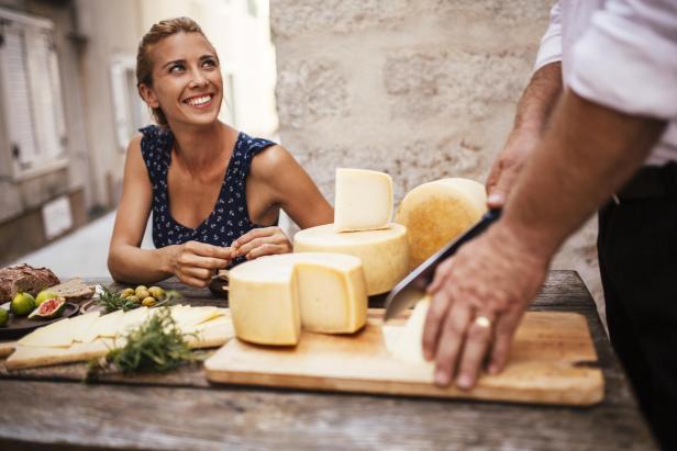 Eine lächelnde Frau sitzt an einem Tisch mit Käse, Brot und Oliven, während ein Mann Käse schneidet.