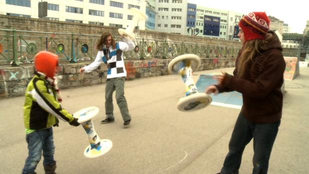 Drei Personen spielen mit Kreiseln auf einer städtischen Freifläche.