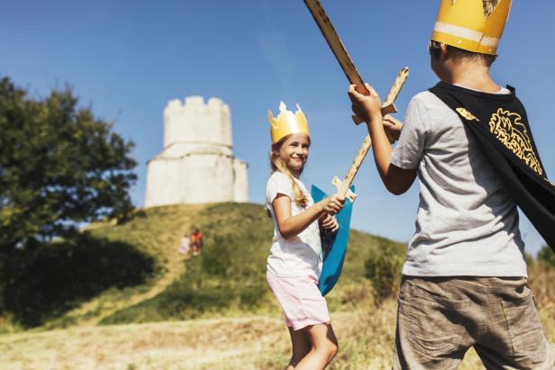 Zwei Kinder mit Kronen und Schwertern spielen vor einer alten Festung.