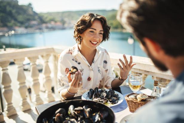 Eine Frau genießt bei einem Mittagessen im Freien Muscheln mit Blick auf das Wasser.