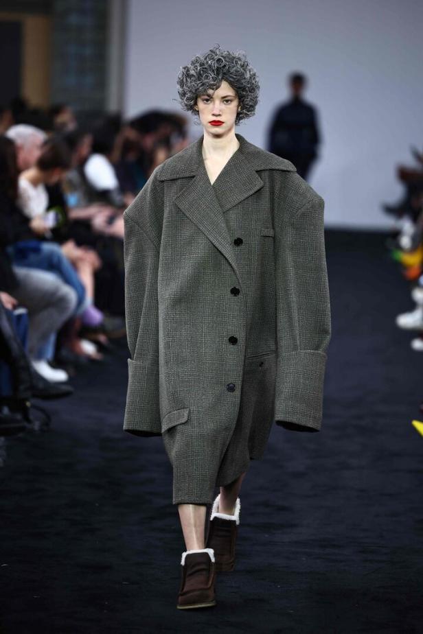 Ein Model mit grauer Perücke und übergroßem Mantel auf einem Laufsteg.