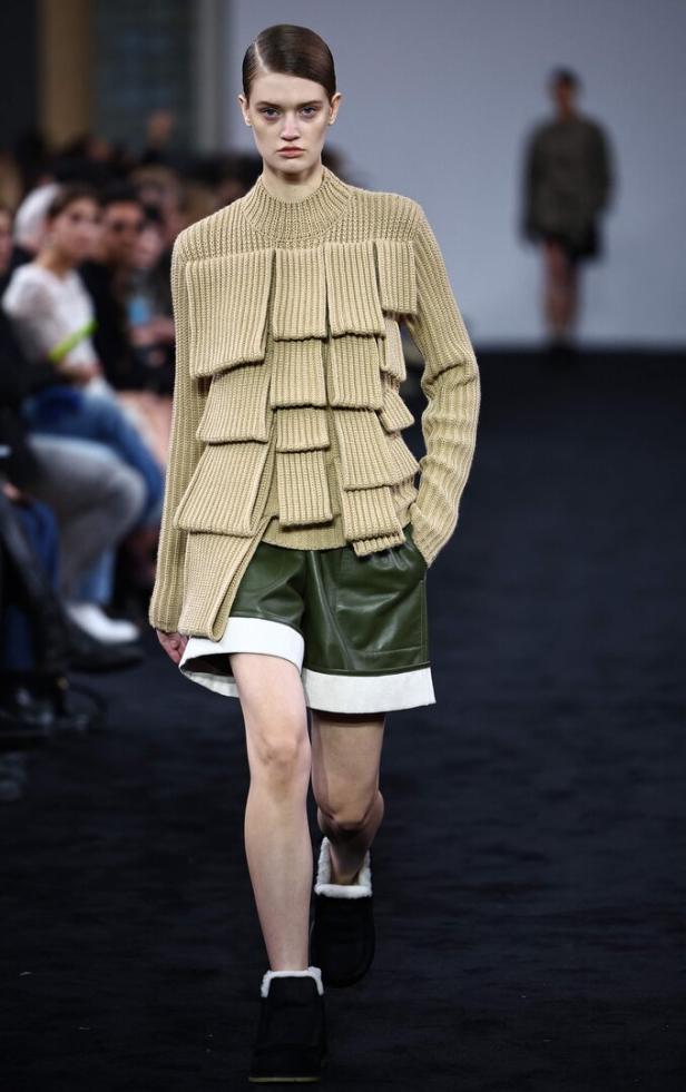 Eine Frau auf dem Laufsteg trägt einen beigen Pullover mit Applikationen und grüne Shorts.