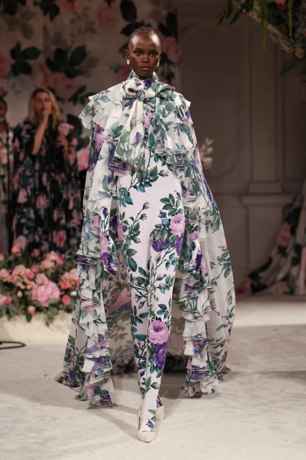 Ein Model präsentiert ein weißes Outfit mit Blumenmuster und Rüschen auf einem Laufsteg.