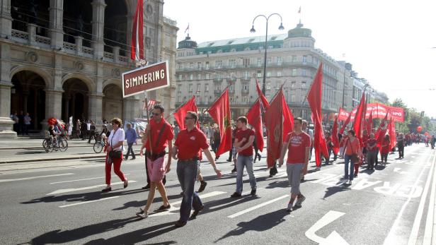 Eine Demonstration mit roten Fahnen zieht durch Wien, erkennbar am Burgtheater.