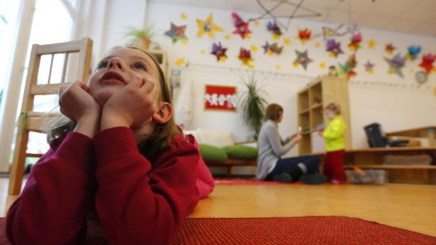 Ein Mädchen liegt auf einem roten Teppich und schaut nach oben, im Hintergrund ein Kindergartenraum.