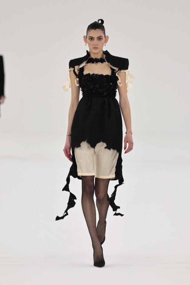 Eine Frau präsentiert ein schwarzes Kleid mit Rüschen und einer beigen Unterlage auf einem Laufsteg.