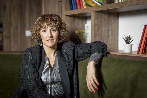 Eine Frau mit lockigem Haar sitzt auf einem grünen Sofa vor einem Bücherregal.