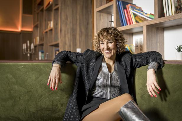 Eine Frau mit lockigem Haar sitzt auf einem grünen Sofa vor einem Bücherregal.