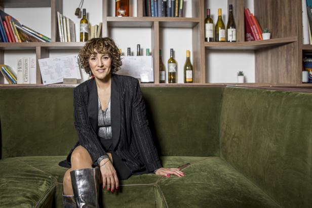 Eine Frau mit lockigem Haar sitzt auf einem grünen Sofa vor einem Bücherregal.