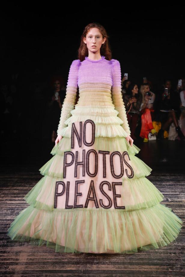 Ein Model trägt ein mehrlagiges Kleid mit der Aufschrift „No Photos Please“ auf dem Laufsteg.