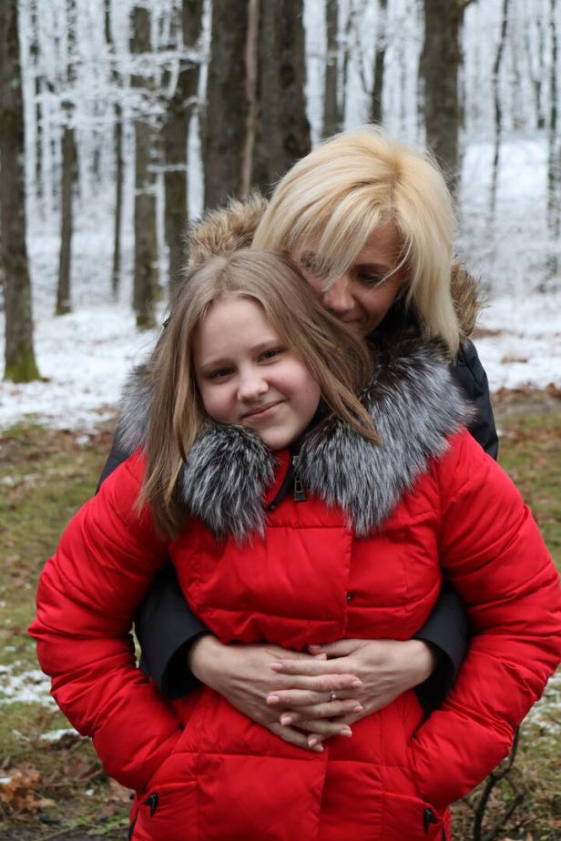 Eine blonde Frau umarmt ein Mädchen mit hellbraunen Haaren und roter Jacke von hinten