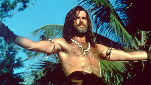Pierce Brosnan als Robinson Crusoe mit ausgebreiteten Armen vor tropischer Vegetation.