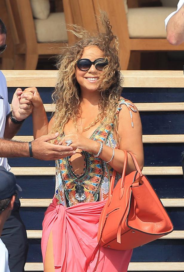 Mariah Carey mit Sonnenbrille, Wickelrock und Handtasche lächelt, während sie Händchen hält.