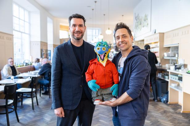 Zwei Männer posieren mit einer bunten Drachenpuppe in einem Café.