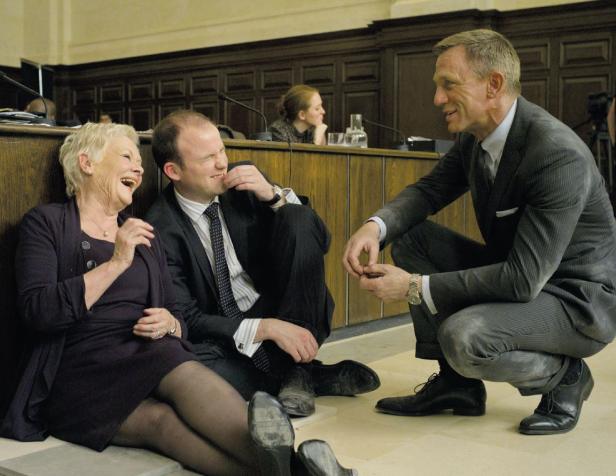 Daniel Craig sitzt kauernd auf dem Boden und unterhält sich mit zwei Kollegen, die lachen.