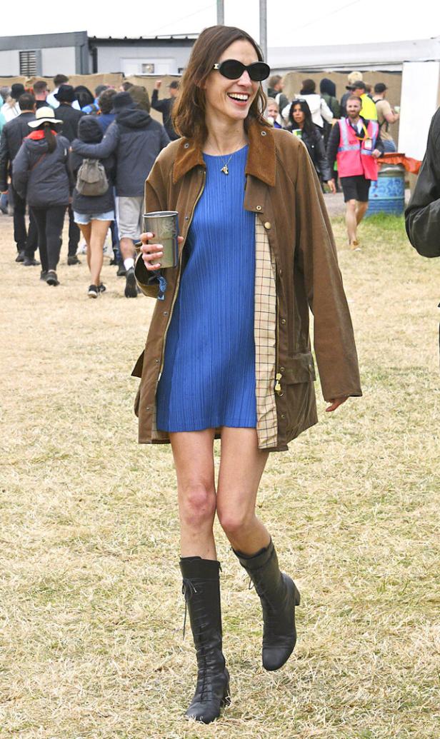 Alexa Chung auf einem Festival, gekleidet in einem blauen Kleid, einer braunen Jacke und schwarzen Stiefeln.