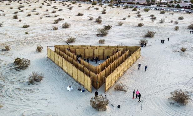 Ein dreieckiges Labyrinth aus Stroh steht in einer Wüstenlandschaft, umgeben von Menschen.