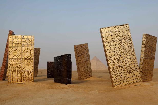 Mehrere dekorative, goldene und schwarze Paneele stehen in der Wüste vor einer Pyramide.