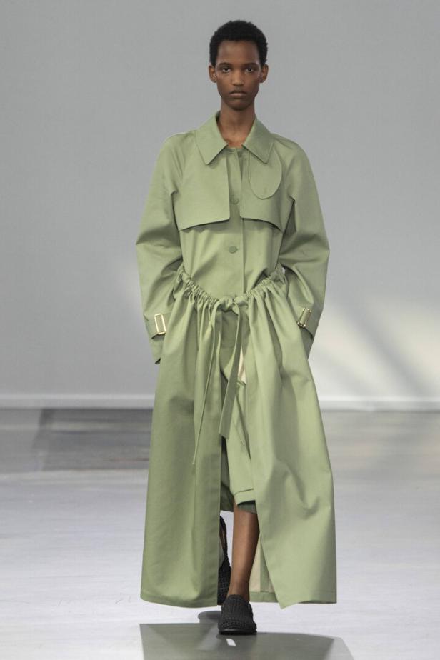 Eine Frau präsentiert einen langen, hellgrünen Trenchcoat mit Kordelzug in der Taille.