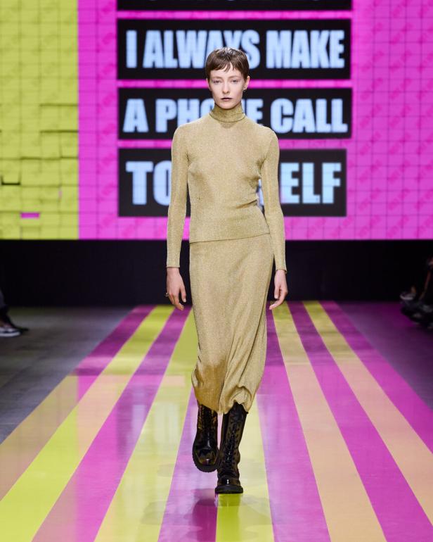 Ein Model in einem goldenen Rollkragenpullover und Rock auf einem Laufsteg.