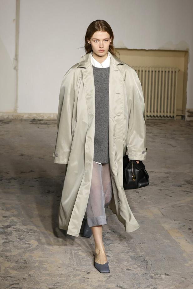 Eine Frau in einem übergroßen Trenchcoat, einem grauen Pullover und transparenten Hosen auf einem Laufsteg.