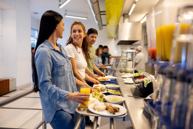 Menschen stehen in einer Cafeteria Schlange, um sich Essen auszuwählen.