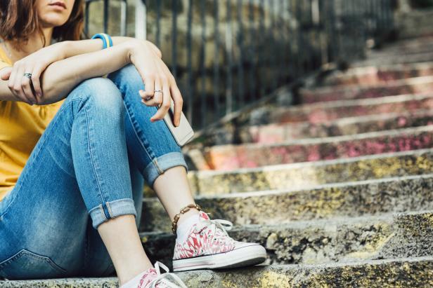 Eine junge Frau sitzt mit Handy auf einer Treppe, gekleidet in Jeans und gelbem Oberteil.