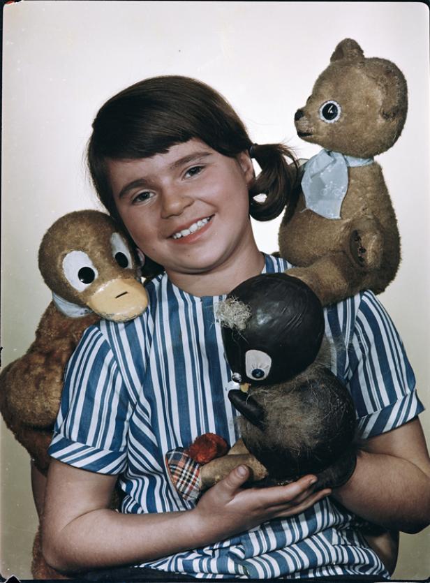 Ein lächelndes Mädchen umarmt mehrere Stofftiere, darunter einen Bären, eine Ente und einen Pinguin.