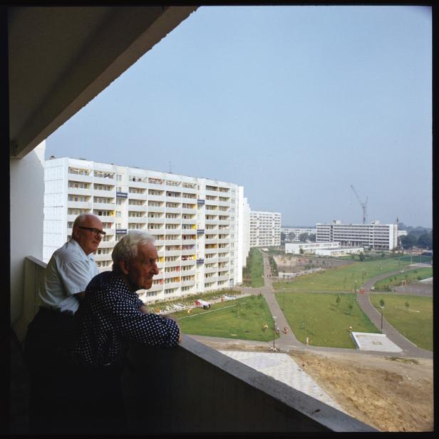 Zwei ältere Männer stehen auf einem Balkon mit Blick auf eine Plattenbausiedlung.