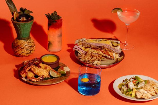 Auf einem orangefarbenen Tisch stehen verschiedene Speisen und Getränke, darunter ein kubanisches Sandwich und Cocktails.
