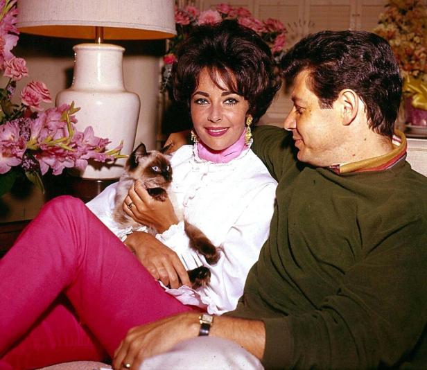 Elizabeth Taylor sitzt mit einem Mann und einer Siamkatze auf einem Sofa.