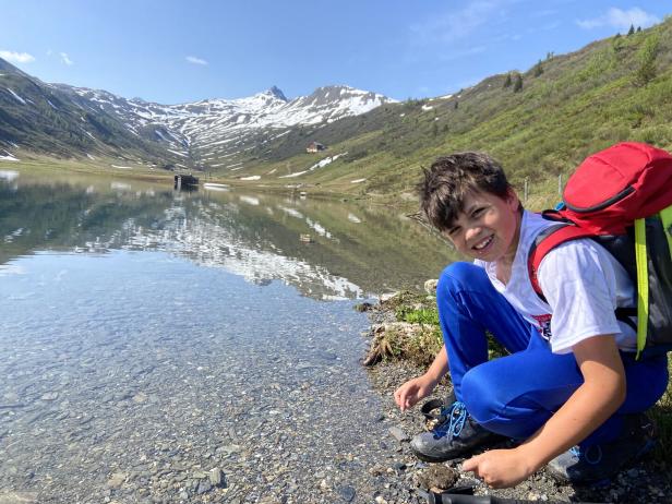 Ein Junge hockt an einem Bergsee mit schneebedeckten Bergen im Hintergrund.