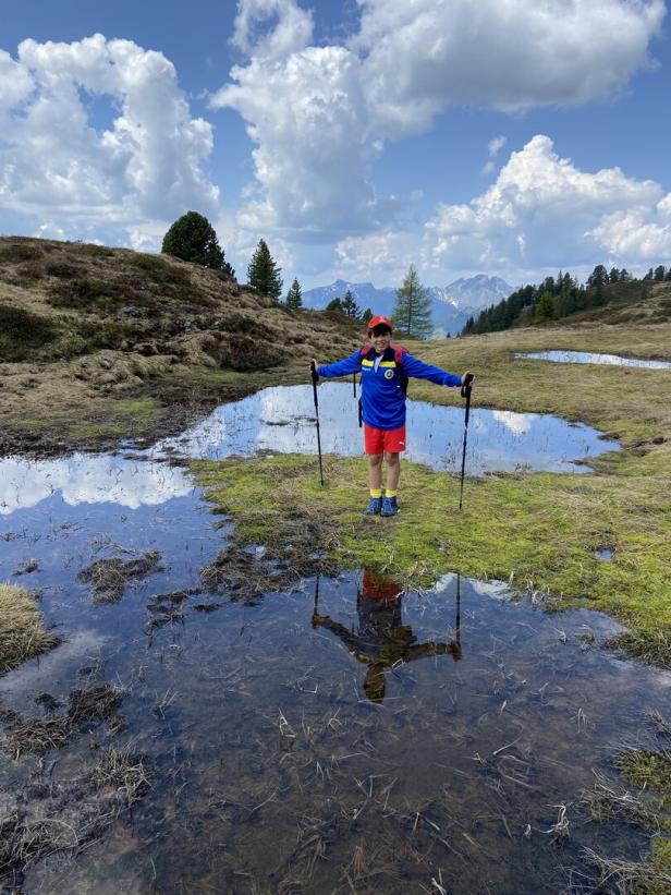 Ein Junge steht mit Wanderstöcken auf einer Wiese mit Wasserstellen und spiegelt sich darin.