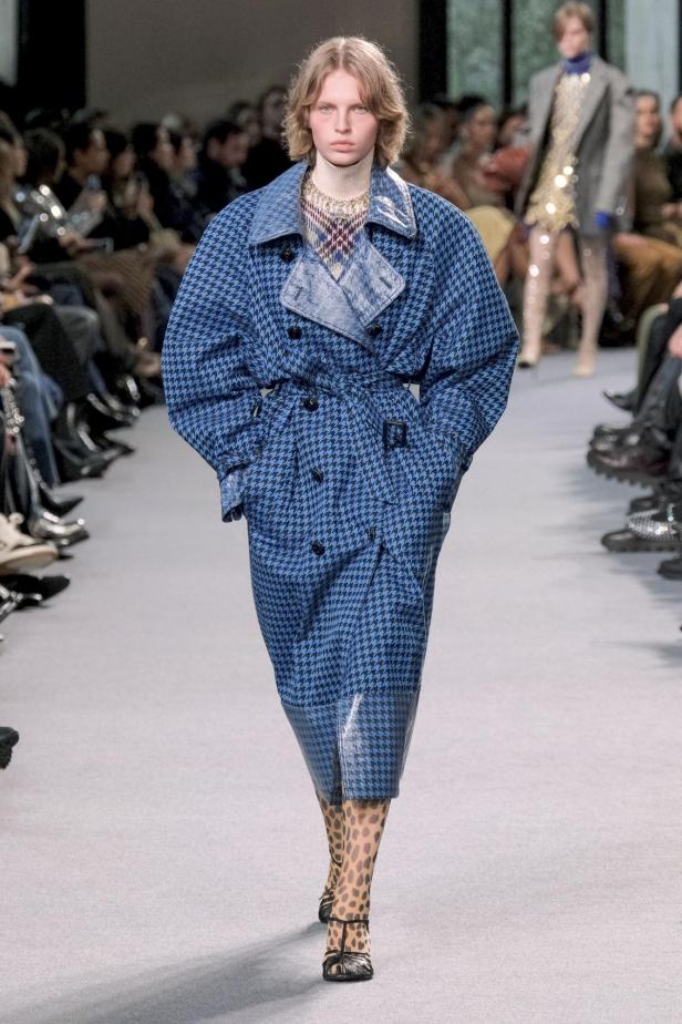 Ein Model präsentiert einen blauen Hahnentritt-Trenchcoat auf dem Laufsteg.