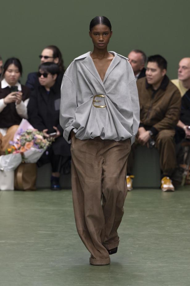 Eine Frau präsentiert ein Outfit mit übergroßer, grau-beiger Bluse und braunen Hosen auf dem Laufsteg.