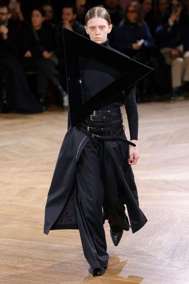 Ein Model präsentiert ein schwarzes Outfit mit einem großen, dreieckigen Accessoire auf dem Laufsteg.