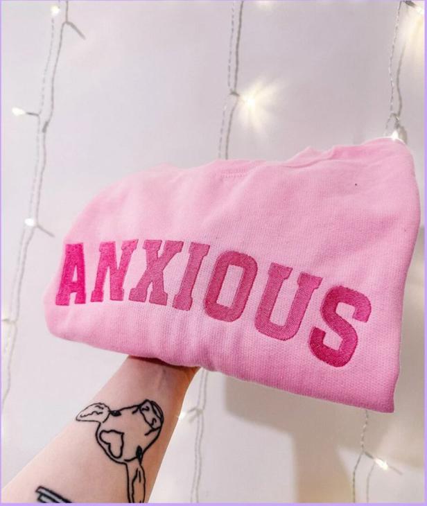 Ein rosa Pullover mit dem Wort „ANXIOUS“ wird von einer Hand gehalten.
